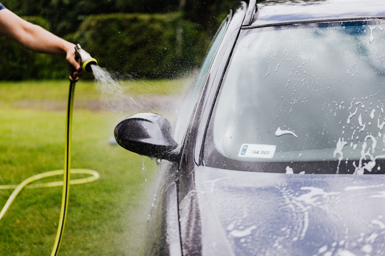 services-05 Person washing a car with a hose outdoors, creating suds on a sunny day.