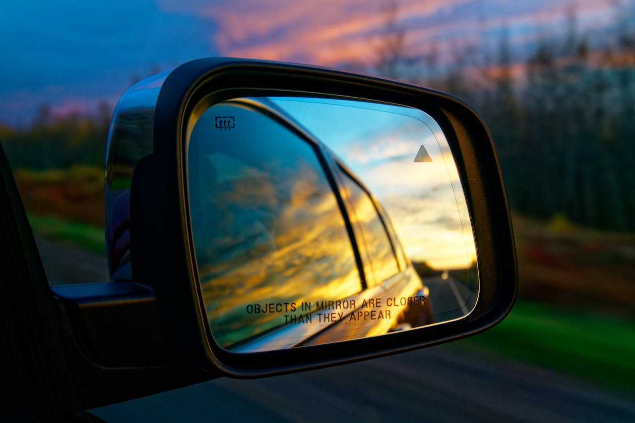 about-03 A car side mirror reflecting a vibrant sunset sky on a highway, creating a colorful and dynamic travel scene.